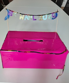 Auf dem Foto ist die Wahlurne für die Betriebsratswahl zu sehen. Der pinke Kasten ist mit  einem Spruchband und farbigen Pailletten verziert
